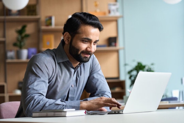 A man at a desk, smiling at his laptop.